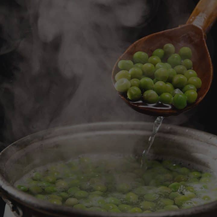A steaming pot of cooked peas with a spoon lifting them out of the water. It has a black overlay.