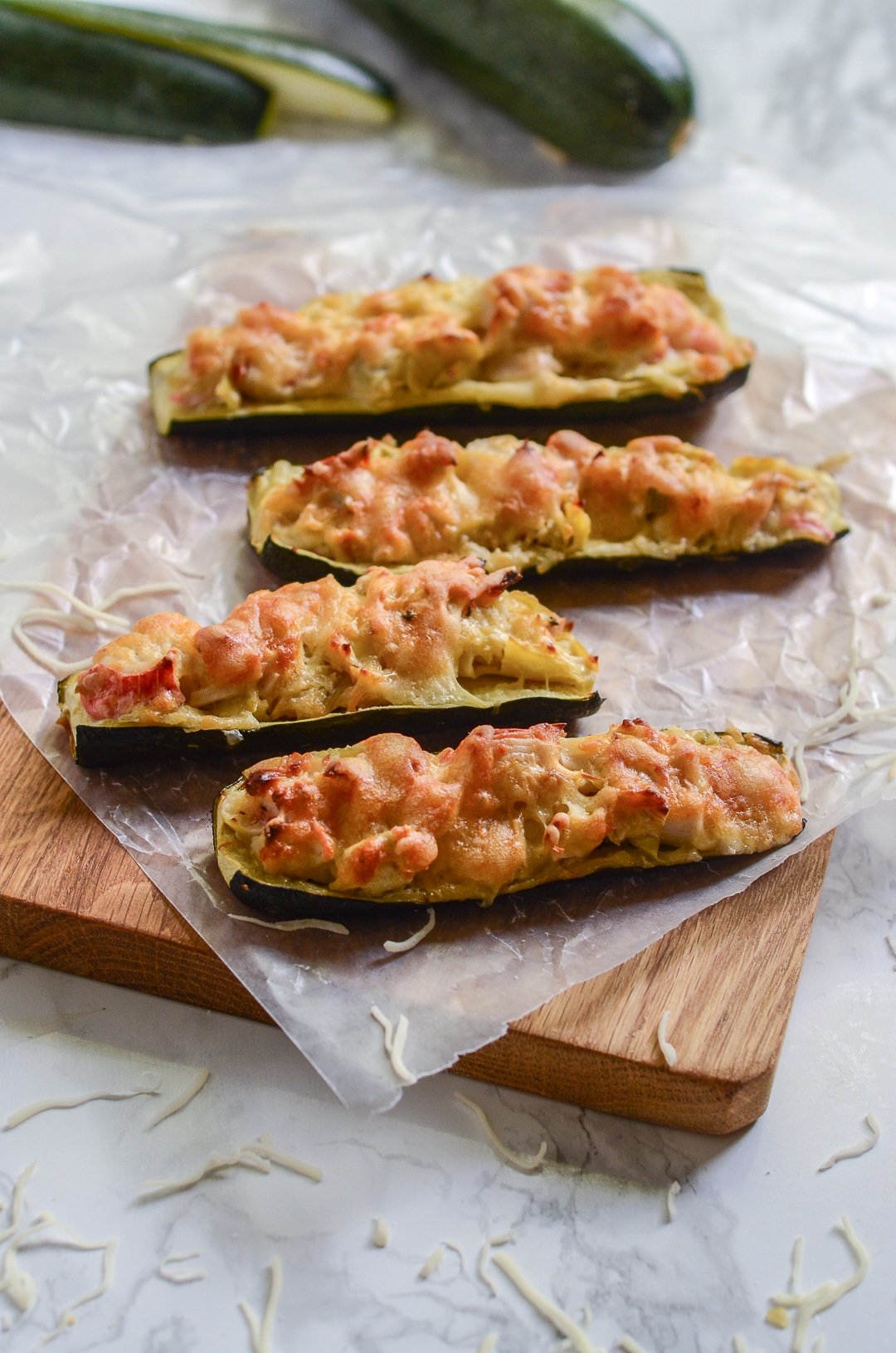Overhead image of four crab and artichoke stuffed zucchini on wax paper and wooden cutting board