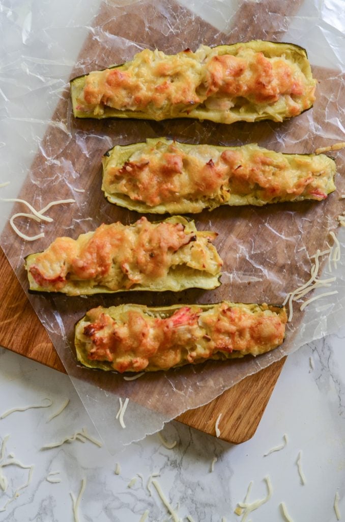 Overhead image of four crab and artichoke stuffed zucchini on wax paper and wooden cutting board