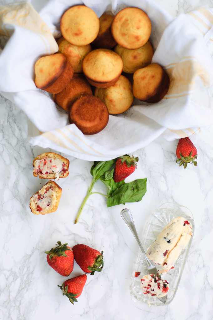 A basket of honey corn muffins next to butter, basil and strawberries.