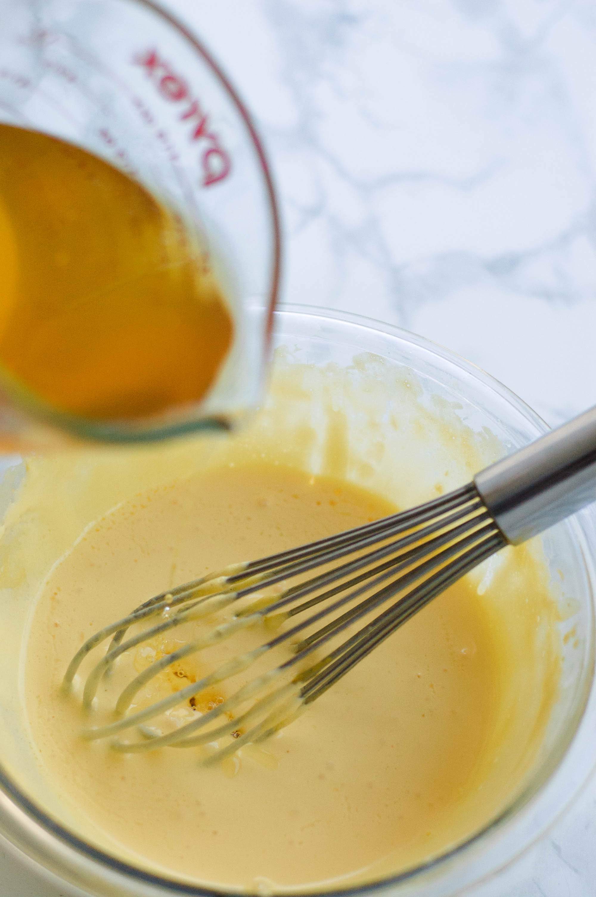 A measuring cup of clarified butter beings poured into a glass bowl of hollandaise sauce with a whisk in it.