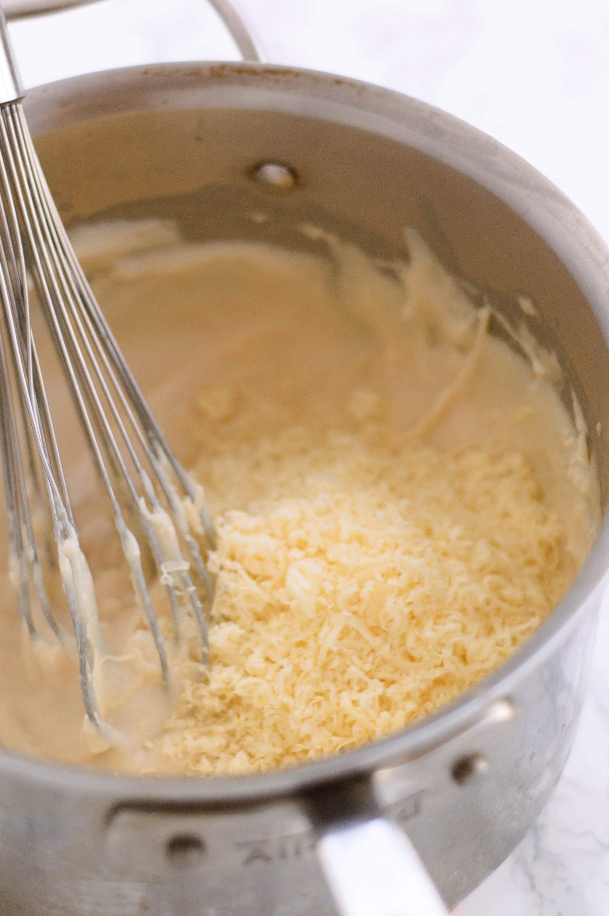 A pot with bechamel sauce and whisk.  Grated cheese is being added in.