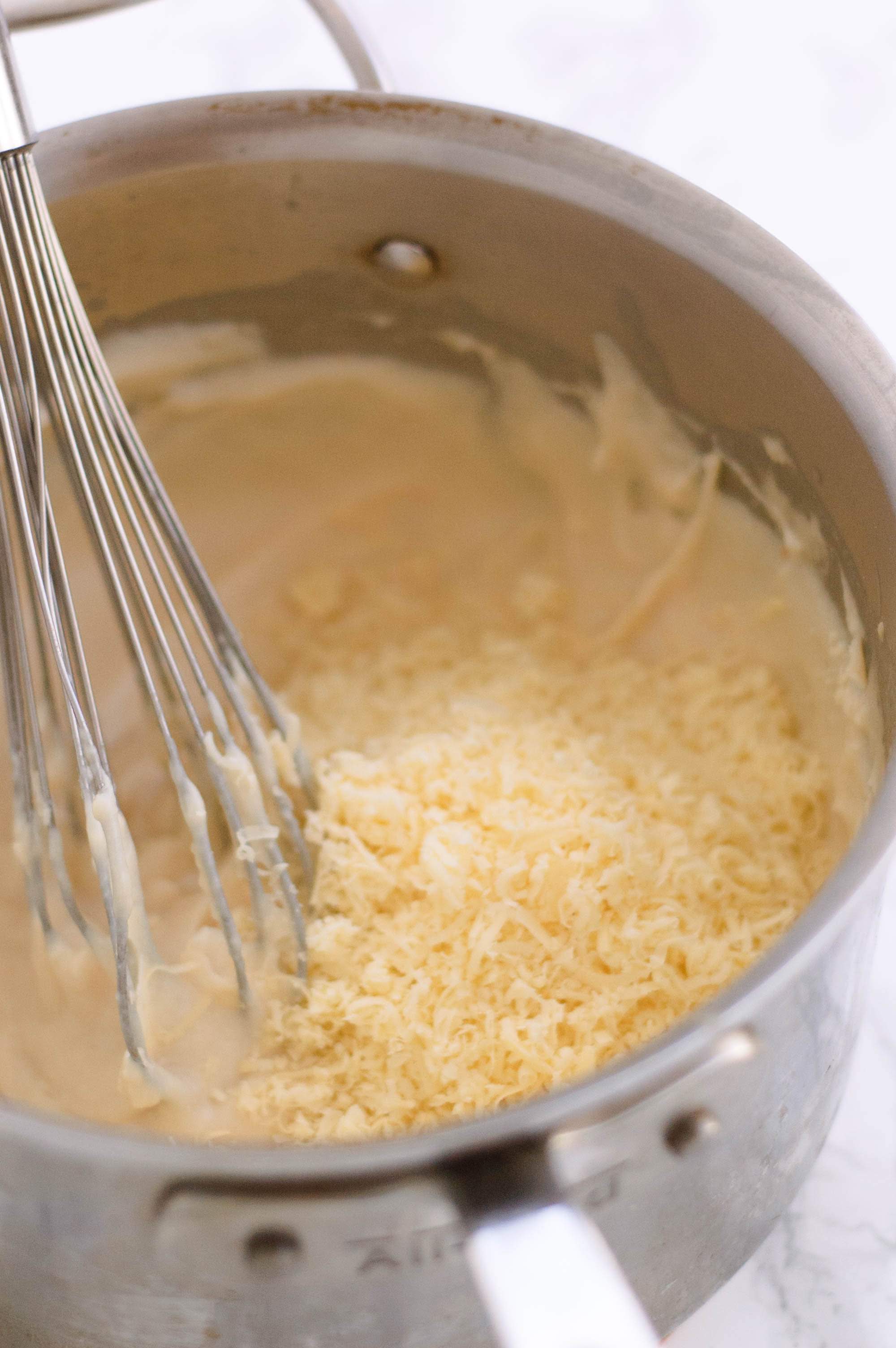 A pot with bechamel sauce and whisk. Grated cheese is being added in.