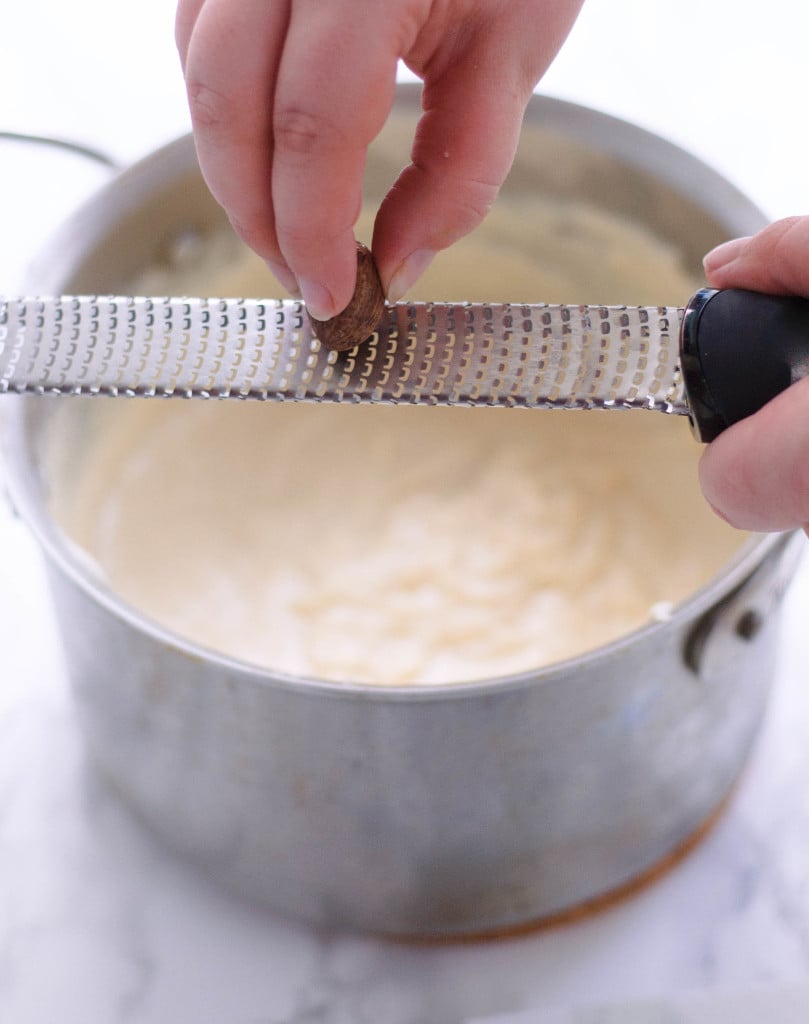 A hand grating nutmeg into a bechamel sauce in a pot.