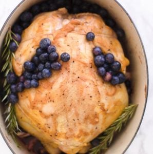 A whole roasted chicken garnished with blueberries and rosemary in a dutch oven.
