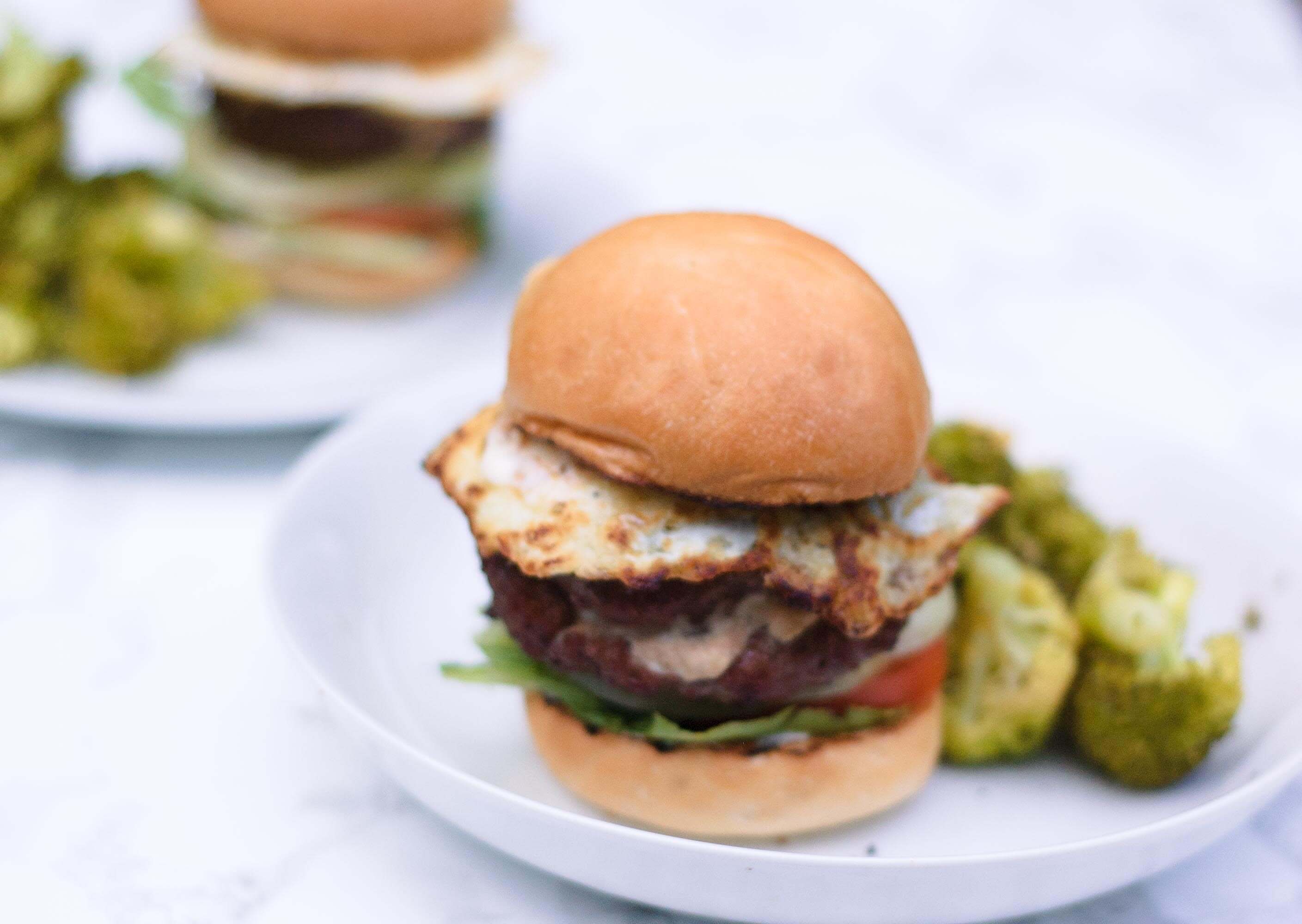 closeup of a breakfast burger stacked high and focused on the fried egg.  It's on a white plate.