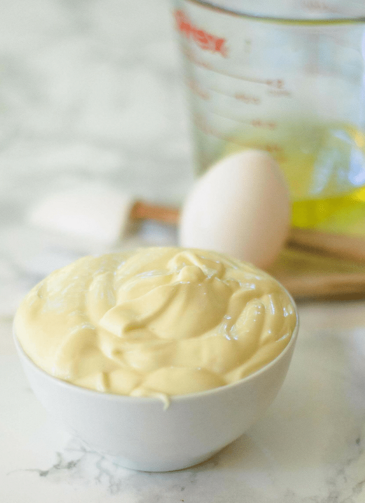 closeup of a small bowl filled with homemade mayonnaise.  Behind it is an egg, oil, and spatula.