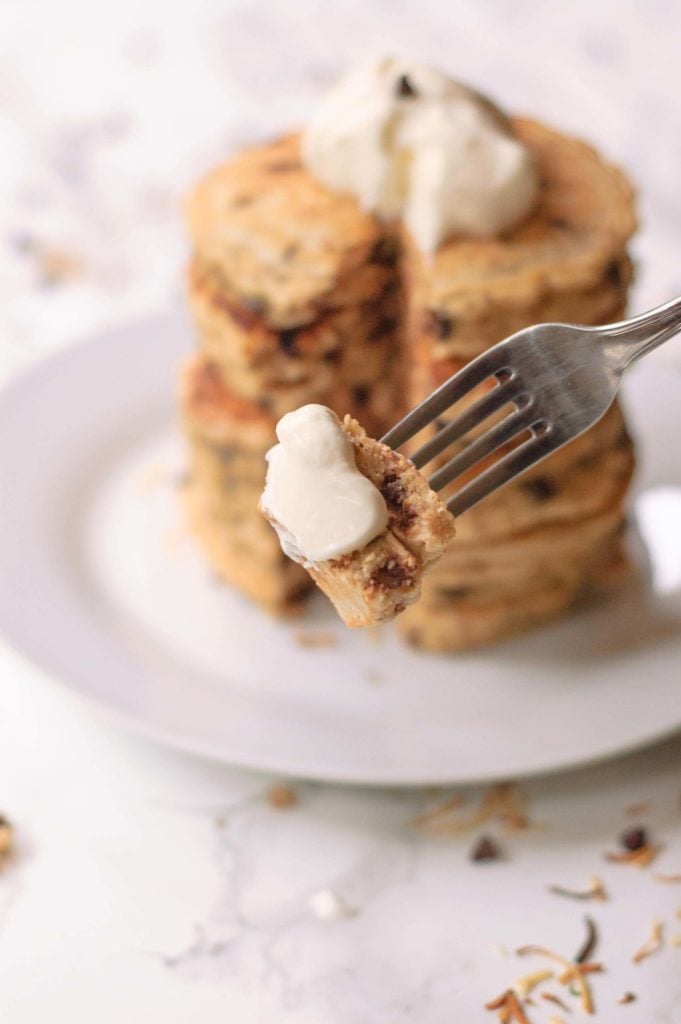 large stack of chocolate chip pancakes in background with a forkful of pancake with whipped cream in foreground