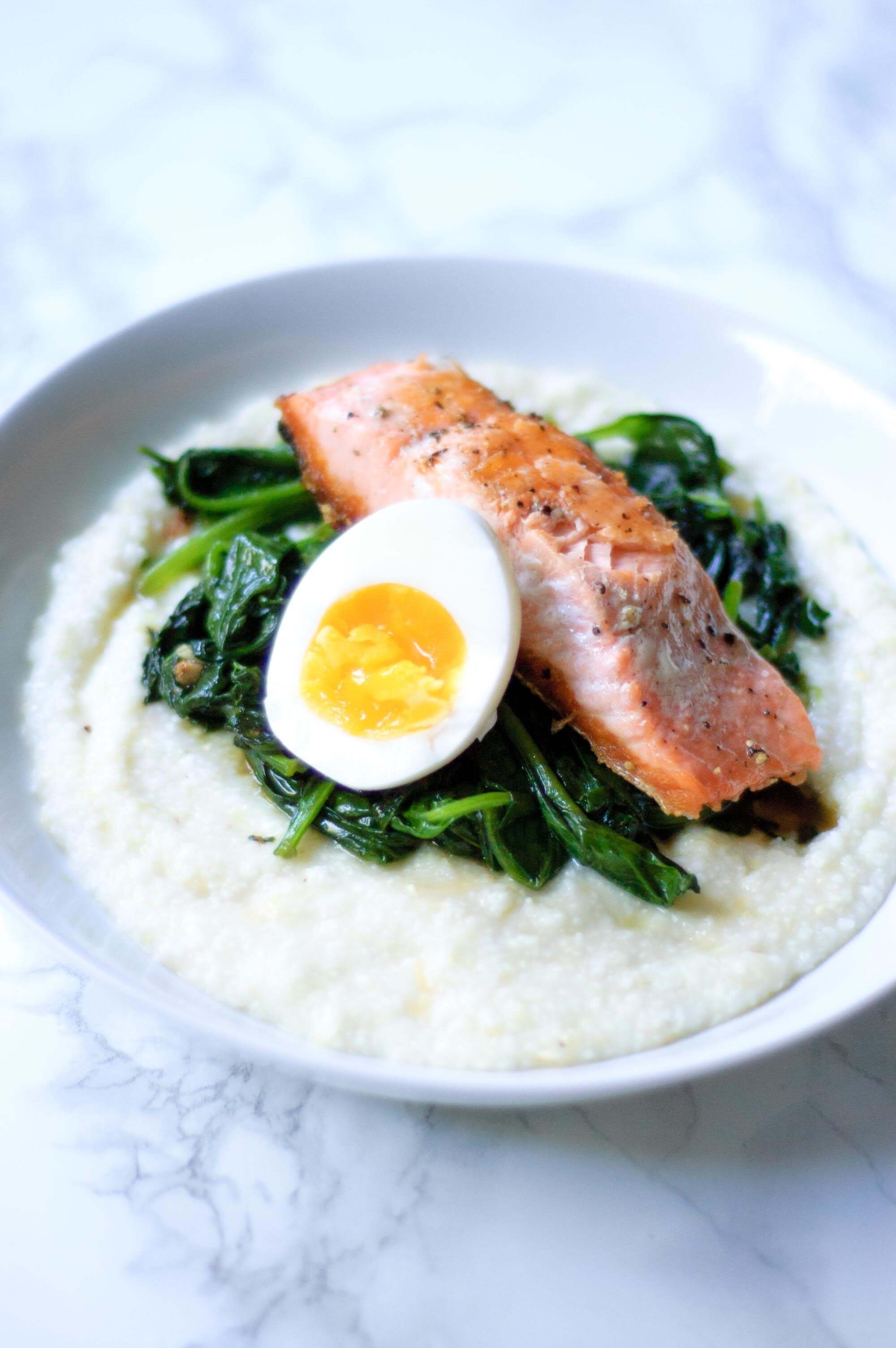 half of a boiled egg next to a serving of salmon with spinach and grits in a white bowl.