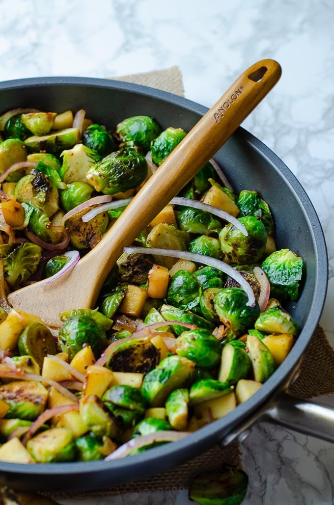 Anolon pan with Brussels Sprouts and apples being stirred with a teak utensil.