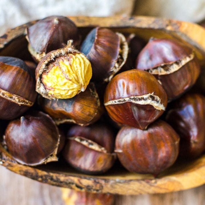 A wooden bowl filled with freshly roasted chestnuts. One is peeled.