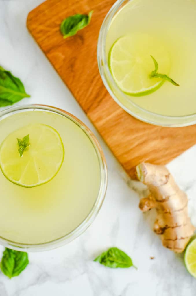 Overhead image of Two cocktail glasses of basil ginger gimlet with ginger, limes, basil and a cocktail shaker