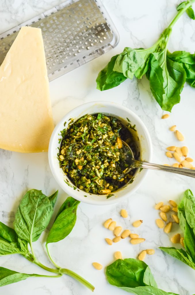 White ramekin filled with pesto surrounded by a wedge of parmesan, pine nuts, and fresh basil