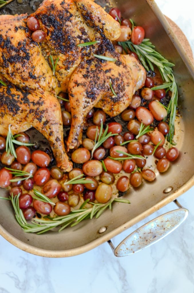 An overhead image of a roasted chicken with rosemary and grapes in a roasting pan