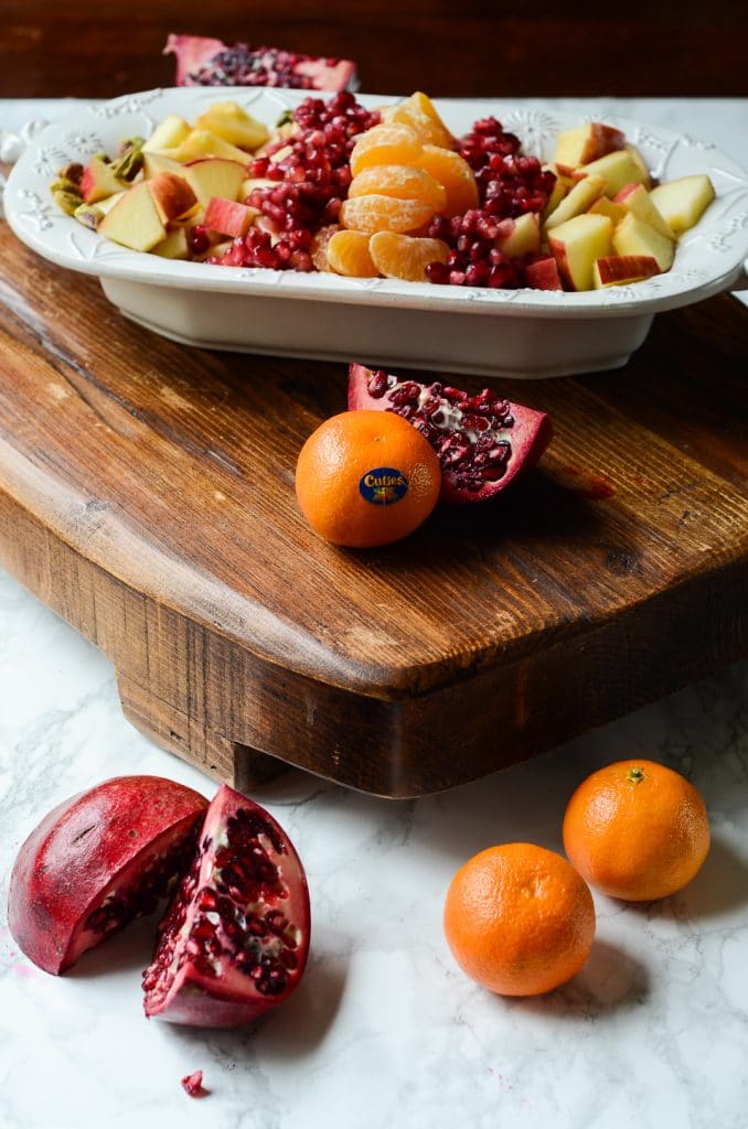 A bowl of winter fruit salad on a wooden board by mandarin oranges and pomegranates.