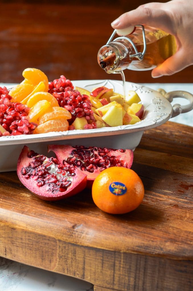 Syrup being poured onto fruit next to a cut pomegranate and cuties mandarin orange.