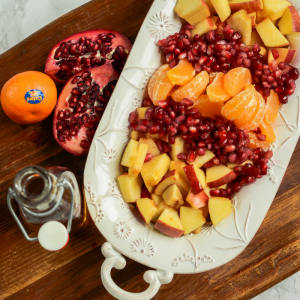 An overhead image of a bowl of winter fruit salad, a container of syrup, pomegranates and Cuties mandarin orange