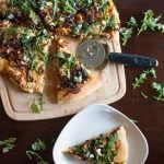 Pizza on cutting board with pizza cutter next to slice of pizza on white plate