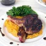 Lamb chops on polenta next to greens on a white plate