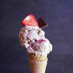 a hand holding a very full sugar cone of ice cream topped with fresh strawberry