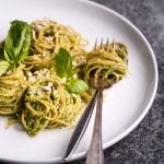 White plate with pasta and basil nexxt to fork and spoon of pasta