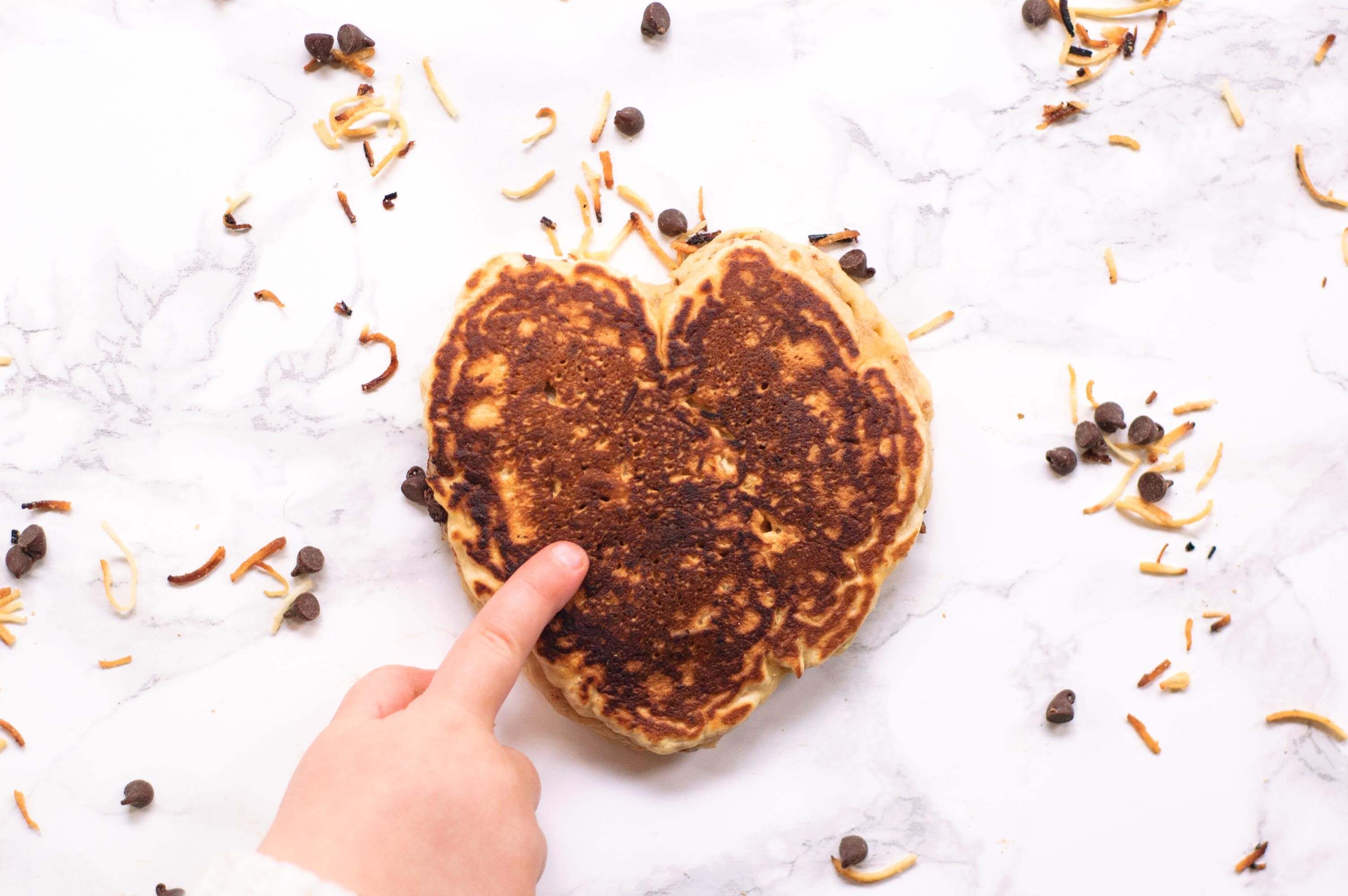 child's finger pointing to a heart shaped chocolate chip pancake