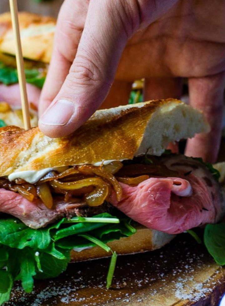 a hand grabbing a prime rib sandwich off of a wooden board
