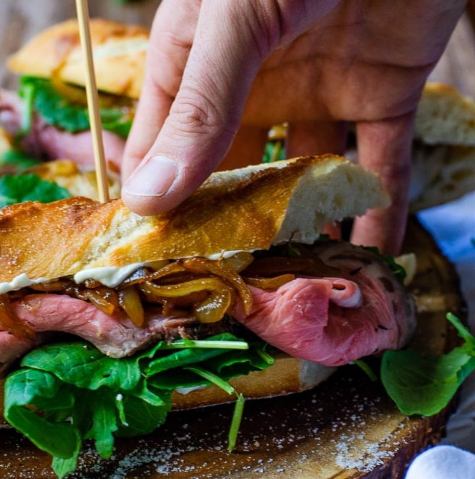 a hand grabbing a prime rib sandwich off of a wooden board