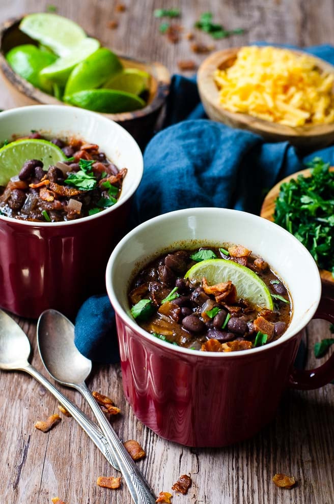 Two red bowls of black bean soup next to bowls of toppings and two spoons