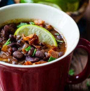 A lime wedge and crispy bacon garnish a red mug of black bean and bacon soup.
