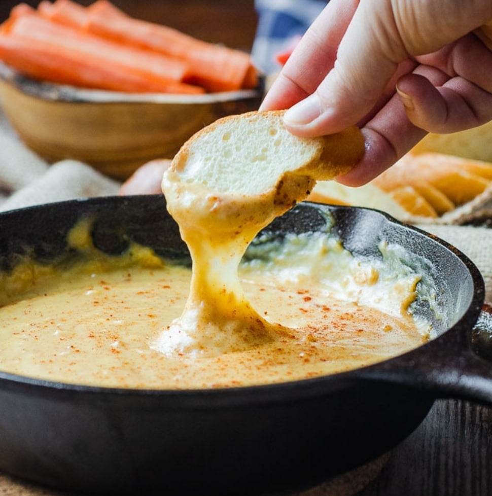 A piece of bread being dipped into smoky beer cheese dip in a cast iron pan
