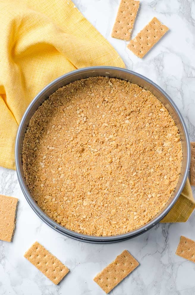 overhead shot of graham cracker crust in a springform pan. It is on top of a yellow napkin and surrounded by graham crackers.