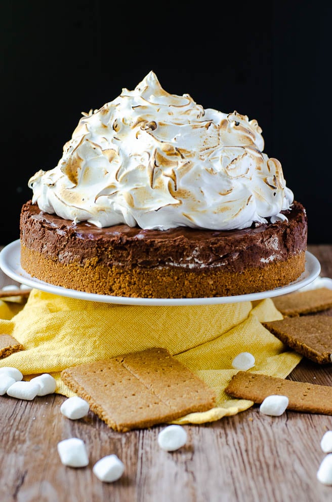 Sky high s'mores pie recipe on a white cake stand. It is surrounded by a yellow napkin, marshmallows, and graham crackers