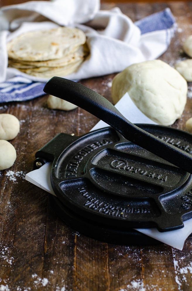 A manual cast iron tortilla press surrounded by fresh corn tortillas and tortilla dough