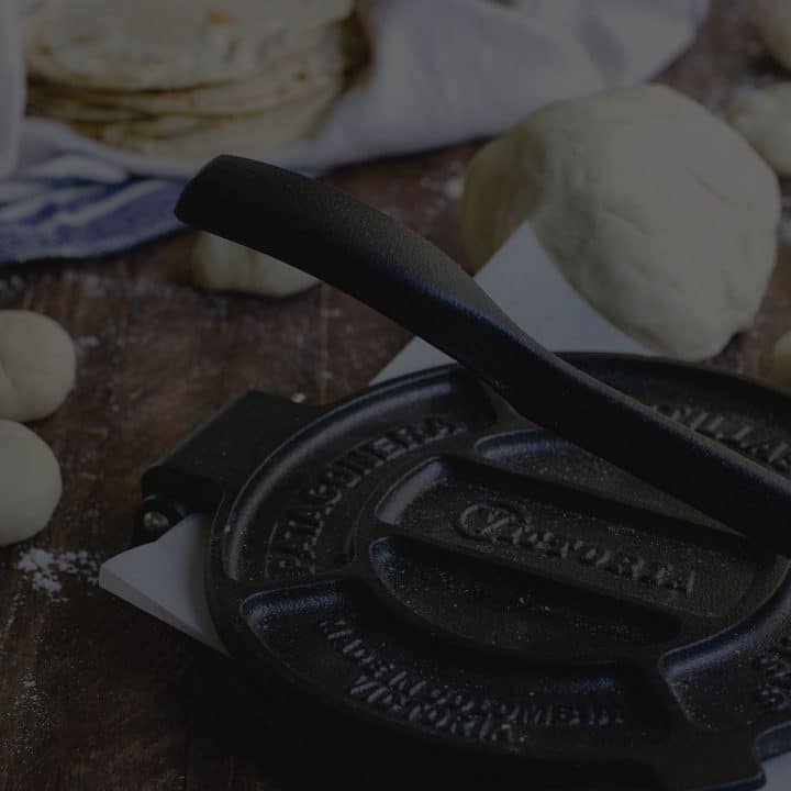 Black overlay on a cast iron tortilla press with fresh tortillas behind it.