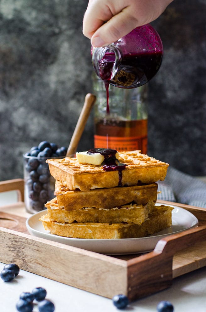 A hand pouring a bottle of blueberry syrup on waffles.