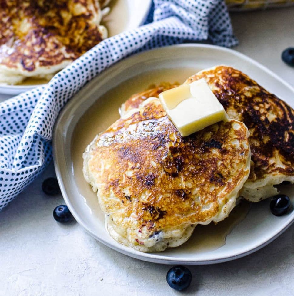 A large pat of butter and syrup on top of fresh blueberry pancakes on a gray plate.