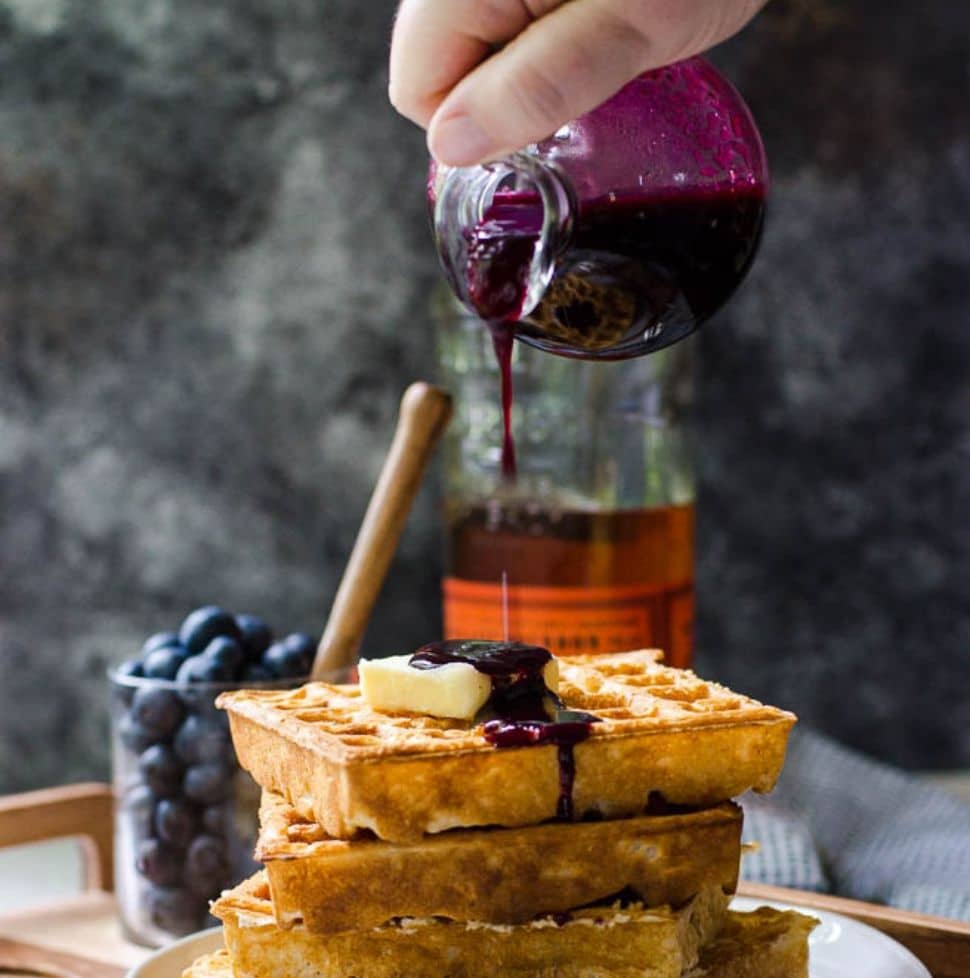 A container pouring blueberry syrup onto a stack of waffles.