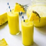 Two glasses of pineapple juice next to a pitcher and a cut pineapple.