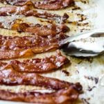 Tongs picking up a piece of bacon cooked on a sheet pan in the oven.