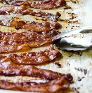 Tongs picking up a piece of bacon cooked on a sheet pan in the oven.