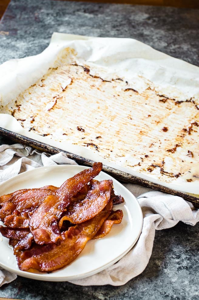 A sheet pan of bacon fat behind a plate of cooked bacon.