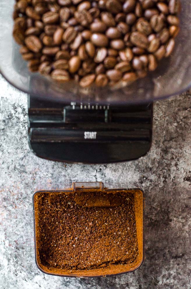 A grinder of coffee beans next to a container of coarsely ground coffee.