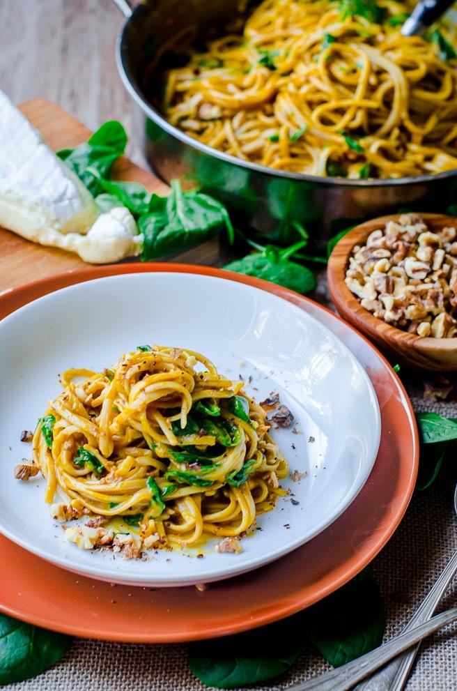 A plate of pumpkin pasta in front of the pan it was cooked in and a wedge of brie.