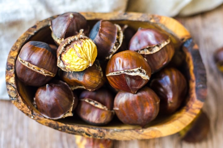 Overhead view of a wooden bowl filled with roasted chestnuts. One is peeled to reveal the nut inside.