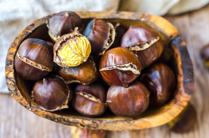 Overhead view of a wooden bowl filled with roasted chestnuts.  One is peeled to reveal the nut inside.