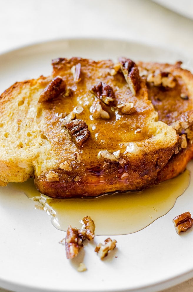 Close up of syrup dripping off of a piece of french toast on a plate.  The french toast has pecans on it.