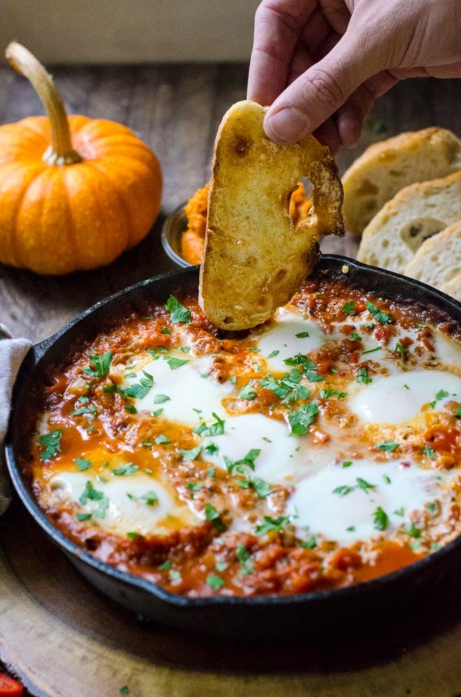 A hand dipping crusty bread into a pan of shakshuka.