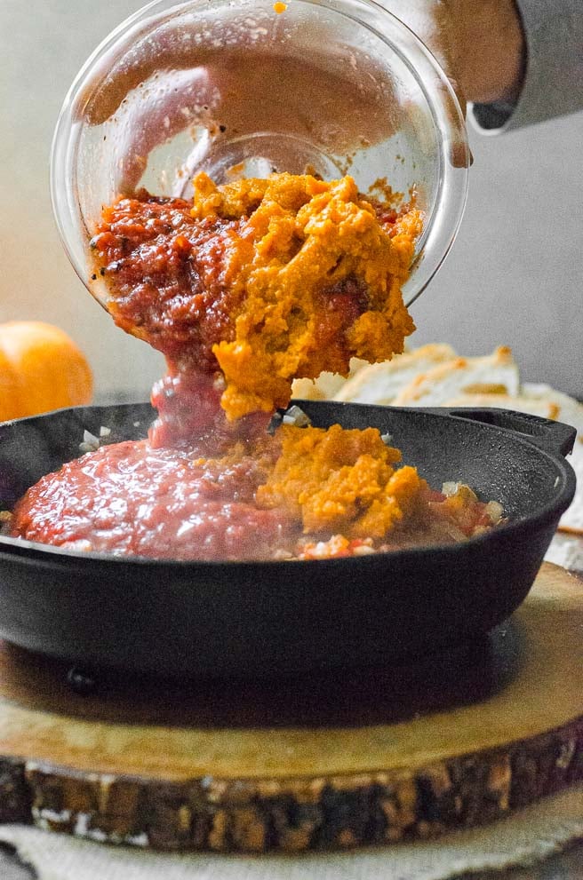 Pumpkin and fire roasted tomatoes being poured out of a bowl and into a cast iron skillet.
