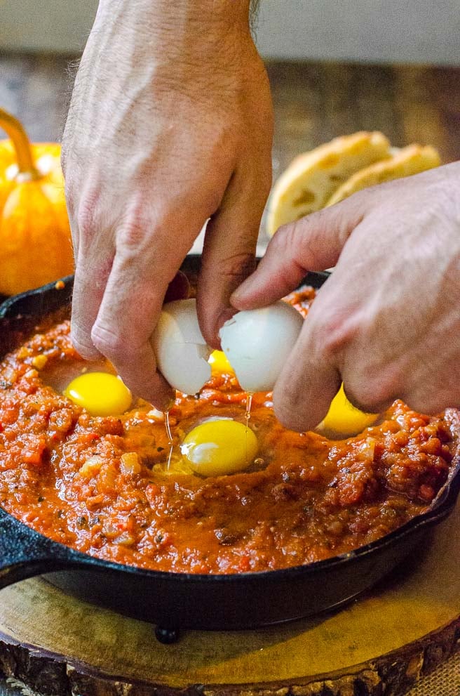 Hands cracking an egg into a pan of tomatoes.